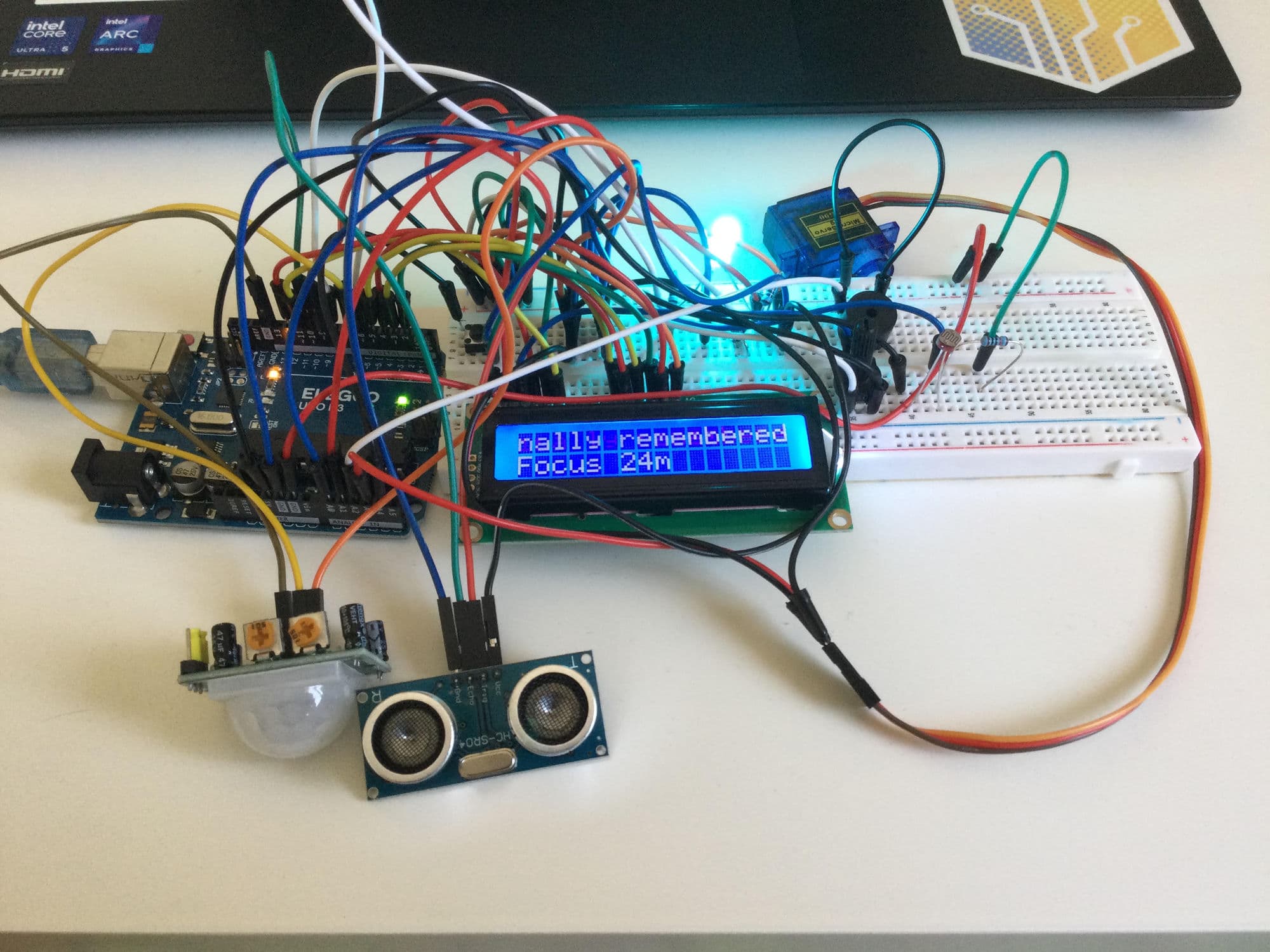 Kivo on a desk: an Elegoo Arduino Uno wired across two breadboards to a blue-backlit 16x2 LCD showing a remembered note and a 24 minute focus timer, with a micro servo, a PIR motion sensor, an HC-SR04 ultrasonic rangefinder and a light sensor wired in beside it.