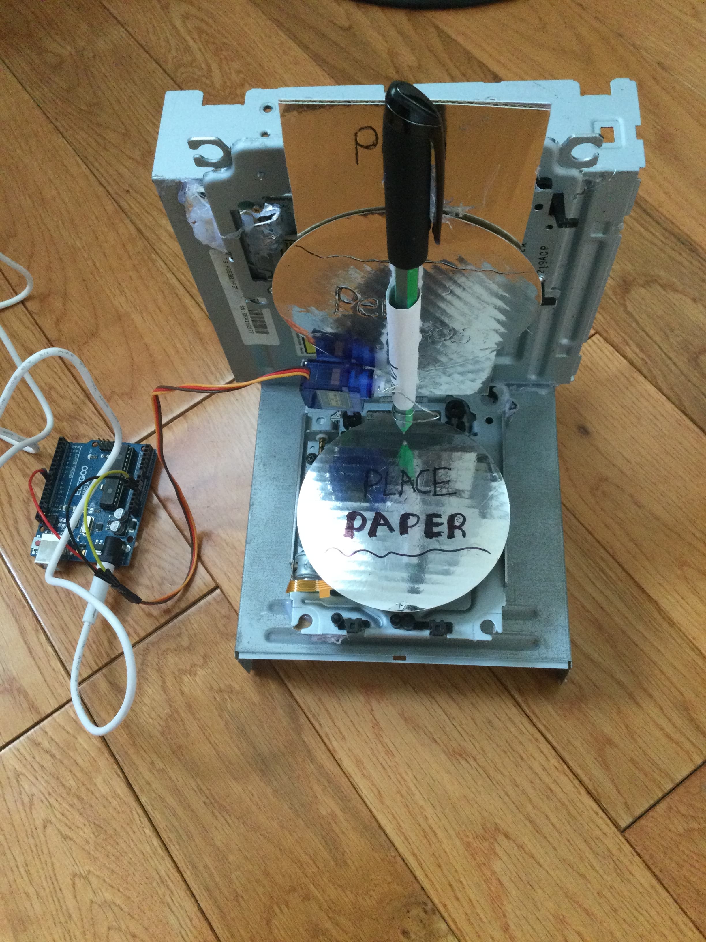 The PenGhost build laid out on a wooden floor: a stripped optical drive chassis with two discs used as platters, a blue micro servo, a marker pen taped to a cardboard arm, PLACE PAPER written by hand on one disc, and an Elegoo Arduino Uno wired in beside it.